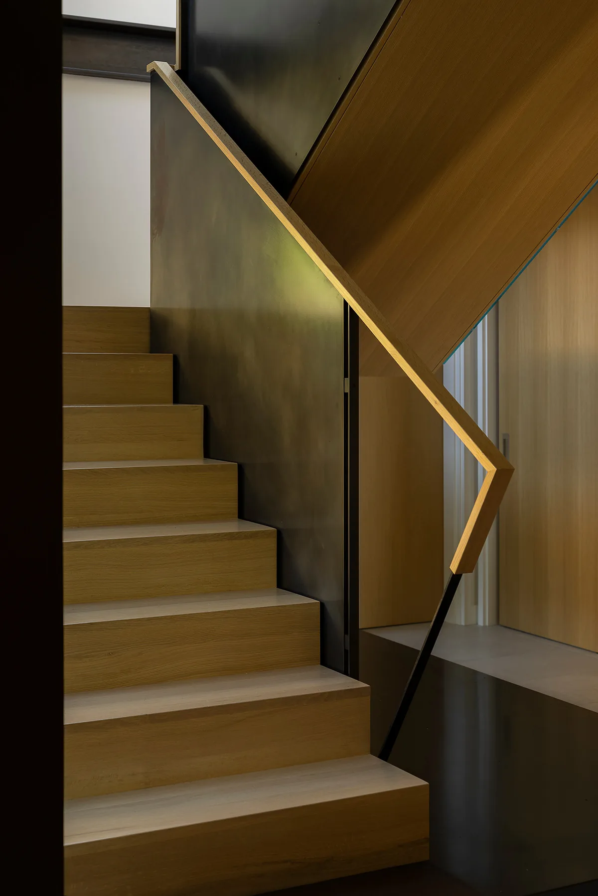 Wooden staircase with minimalist handrail and contrasting dark paneling in a modern interior.
