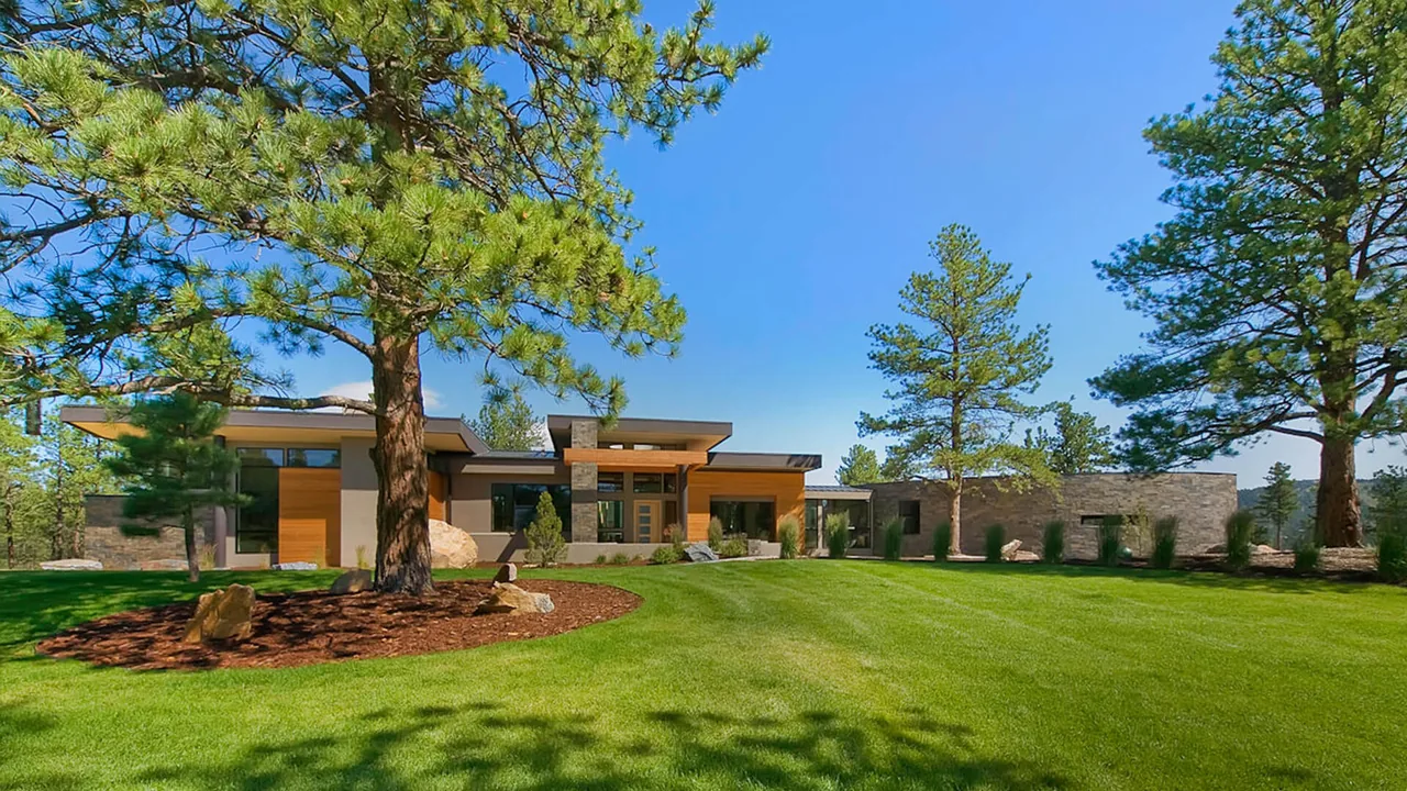 Modern single-story home with flat roofs, wood and stone exterior, situated in a natural landscape with large pine trees.
