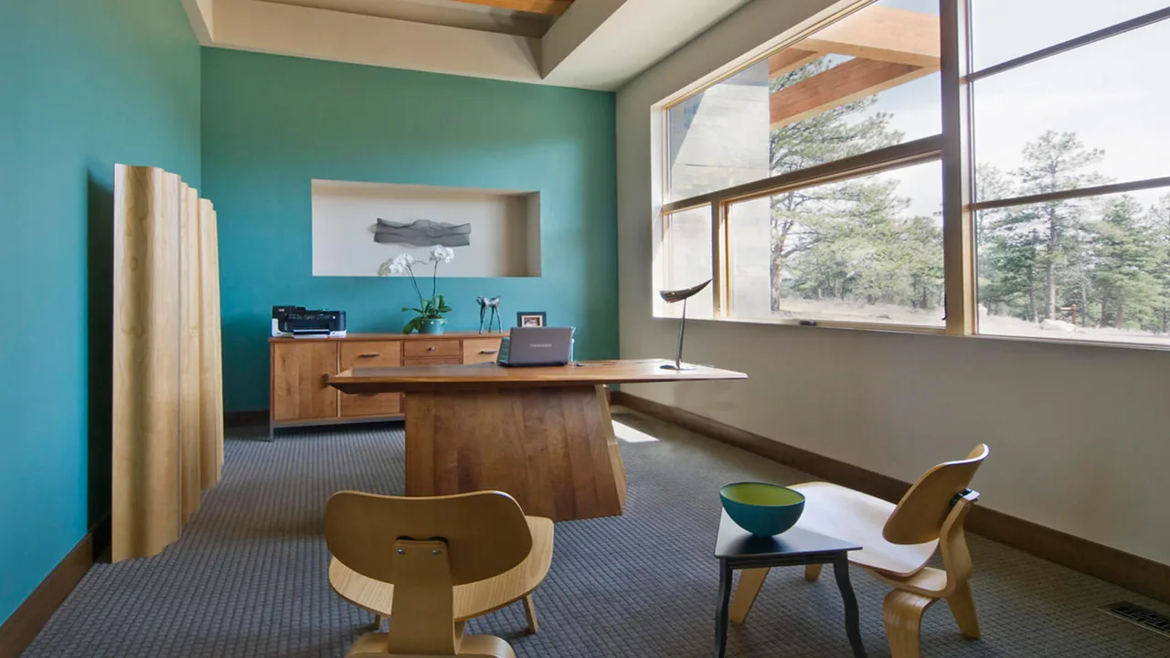 The modern home office features a wooden desk, two chairs, and expansive windows overlooking pine trees.