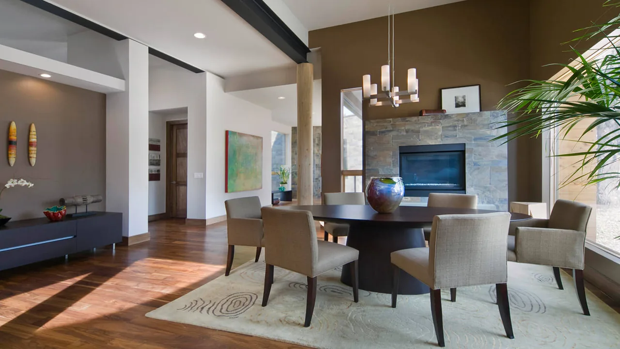 Dining space with a round table and six upholstered chairs next to a stone fireplace and large window.