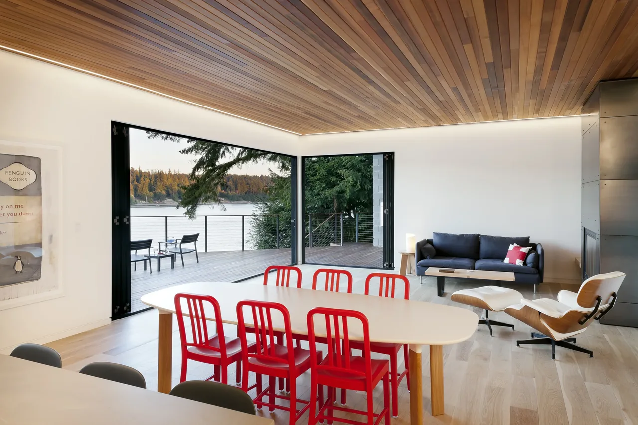 Open-plan dining and living room with large folding glass doors opening to a deck overlooking a lake and forested shoreline.