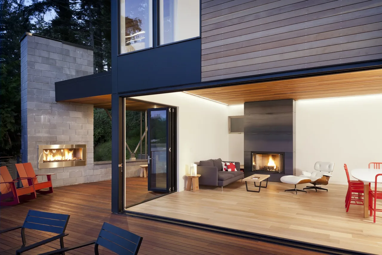 Herron Island Cabin features floor-to-ceiling glass doors that open to a wood deck with an outdoor fireplace and seating.