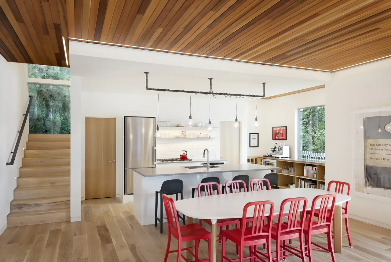 Open-plan kitchen and dining space features light wood flooring, a wood ceiling soffit, and a minimalist staircase.