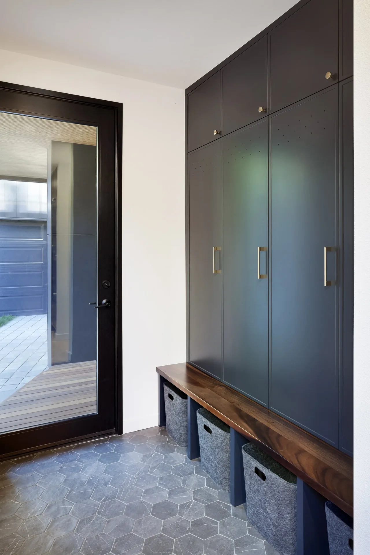 Mudroom features dark gray built-in cabinets with brass handles, a wooden bench with storage bins, and hexagonal tile flooring.