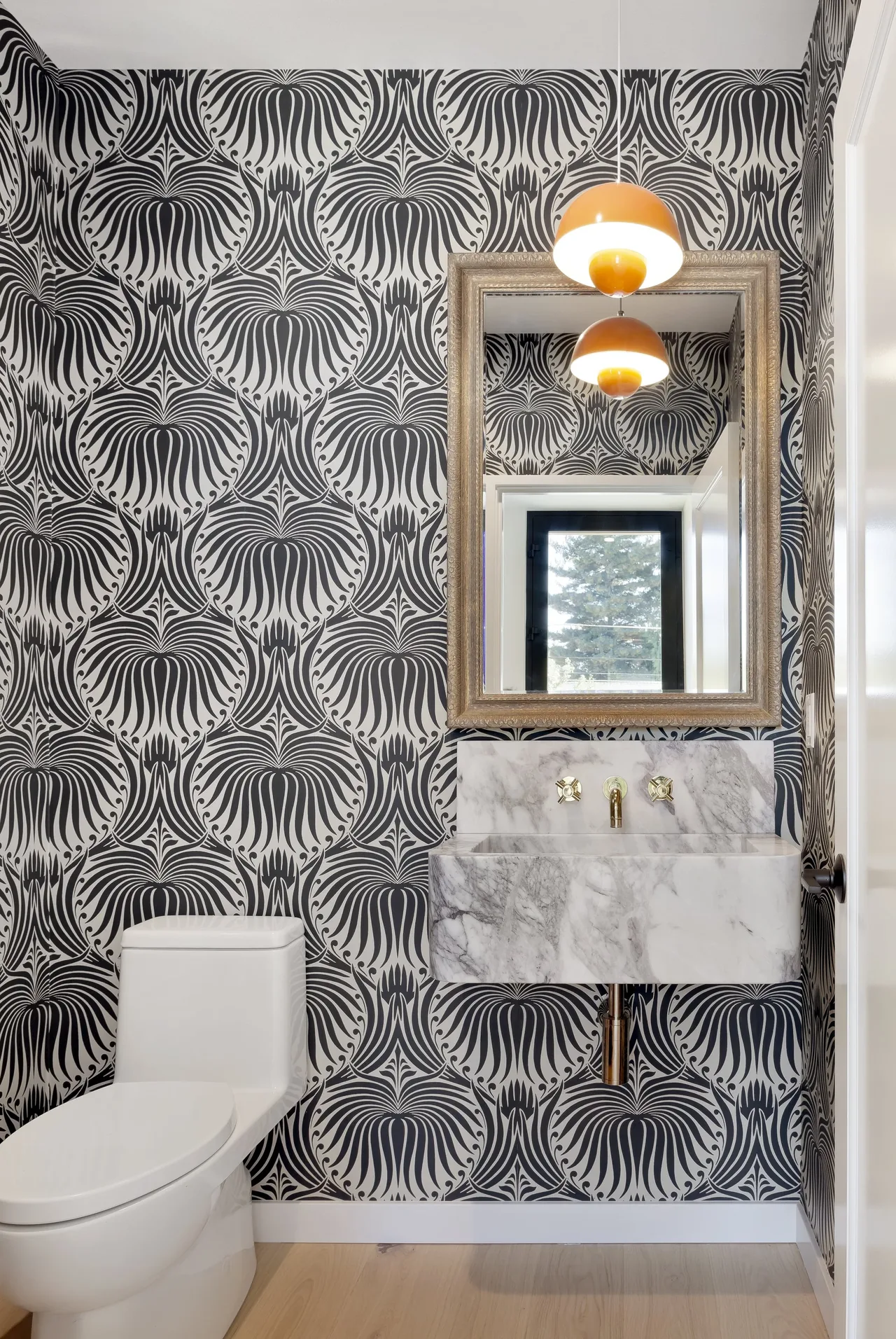 A bathroom featuring monochrome patterned wallpaper, a marble sink with gold fixtures, and a traditional framed mirror.