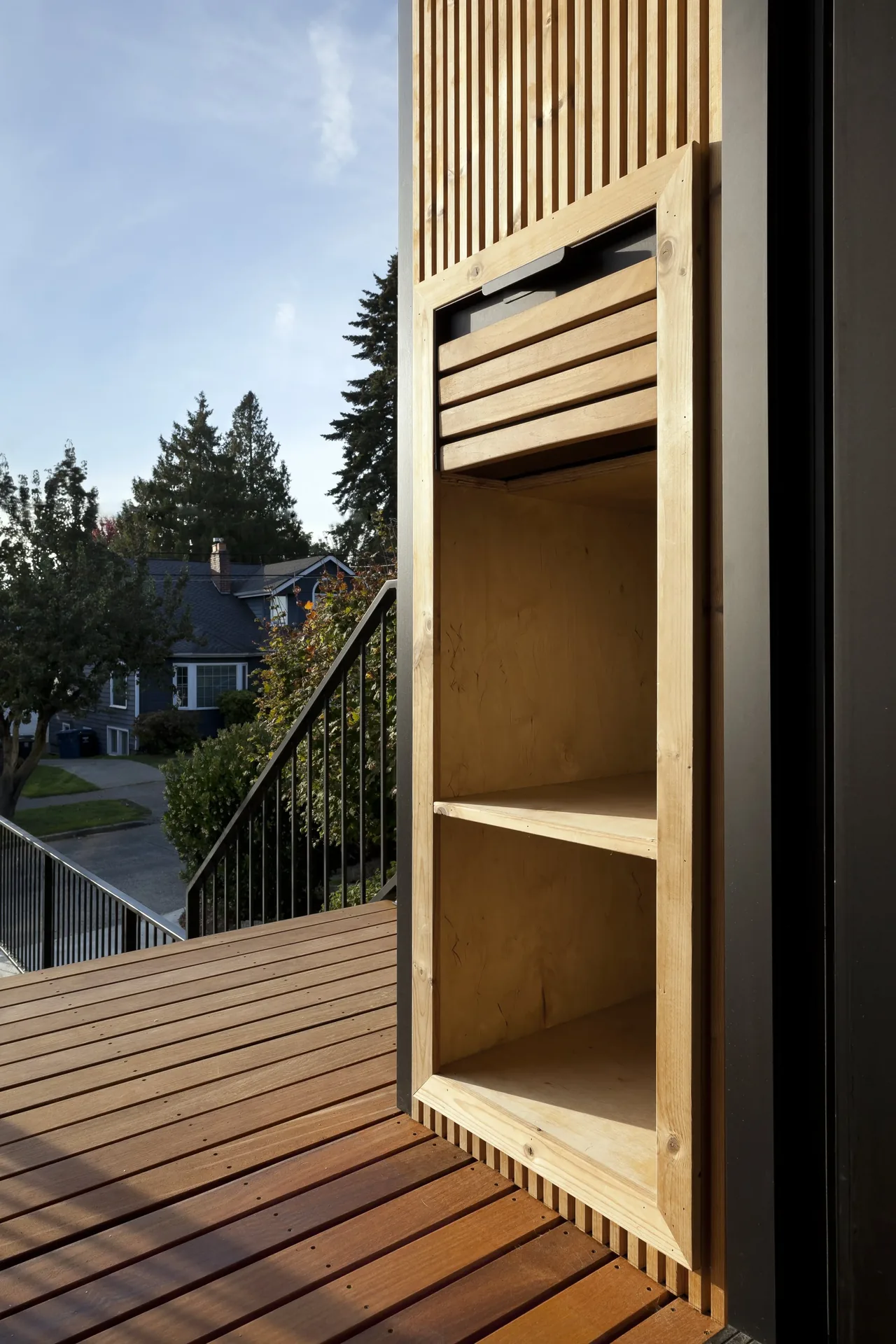 Outdoor wooden shelving integrated into a vertical slat wall on a deck with a view of neighboring houses.