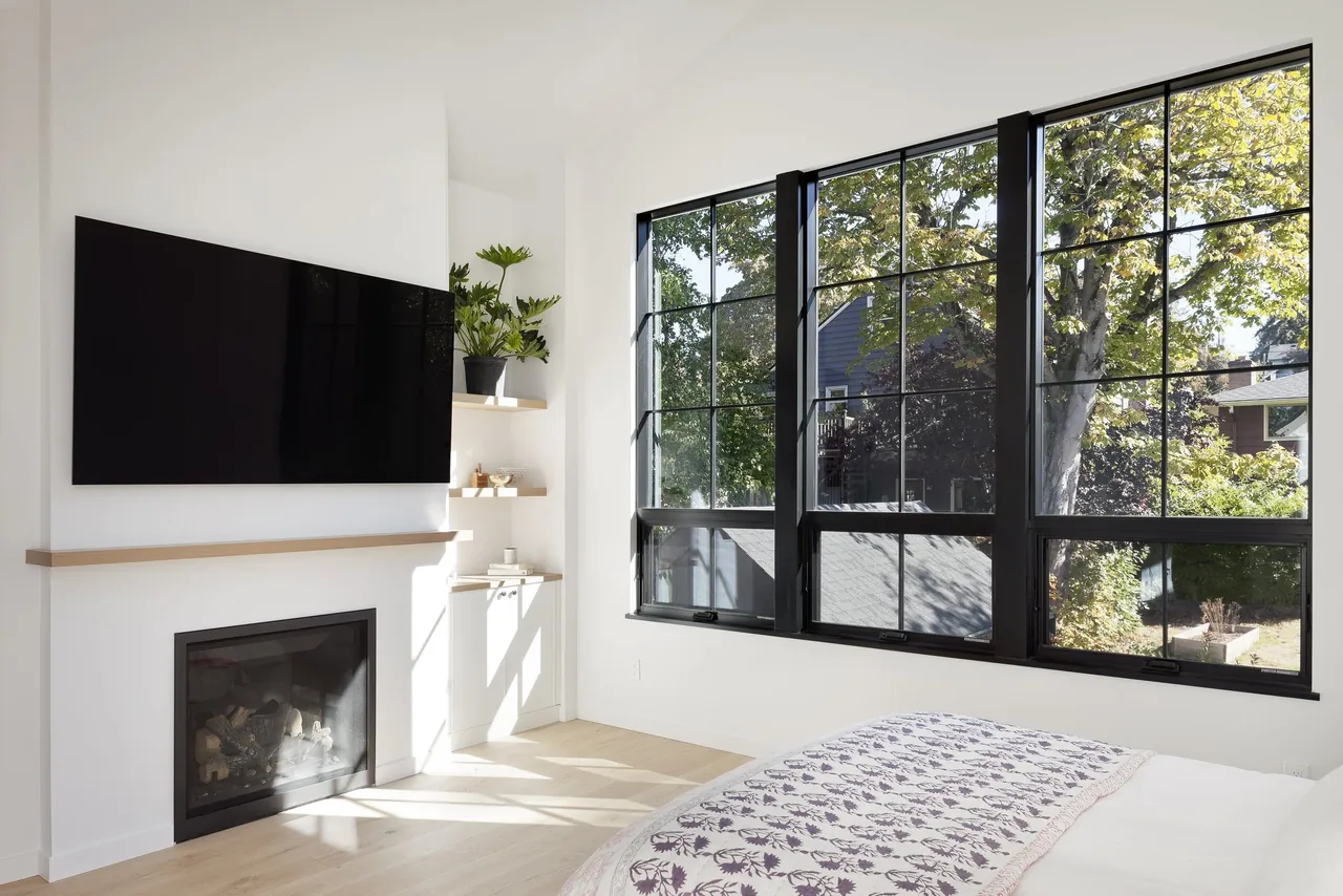 Bedroom corner features large black framed windows, a fireplace under a wood mantel, and a mounted TV above.