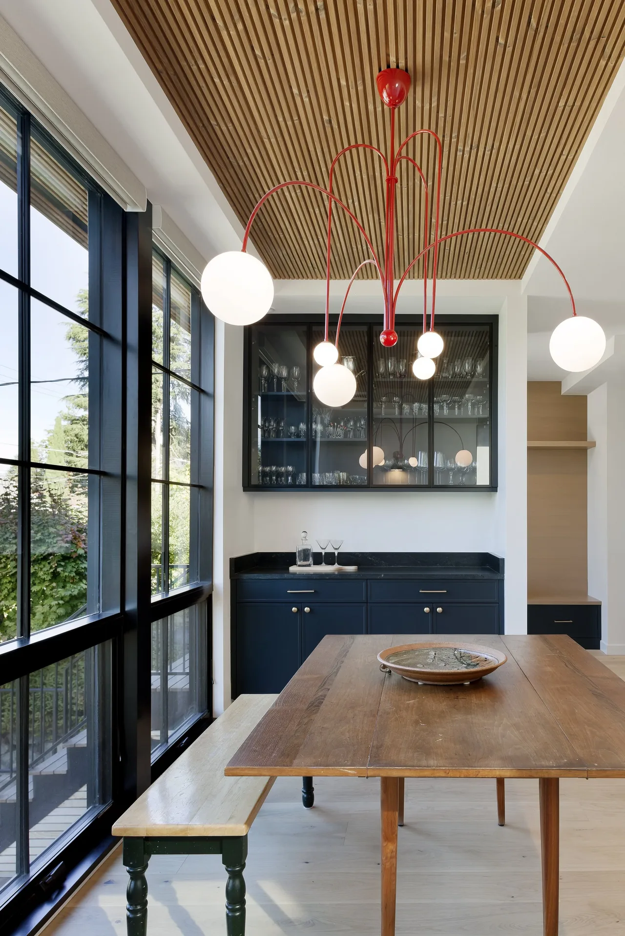 Ravenna House dining area features black-framed grid windows, a red pendant chandelier, and a wood slatted ceiling.