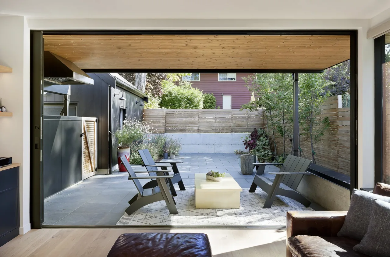 Covered patio seating area with wood slatted ceiling and black trimmed sliding door opening to garden courtyard.