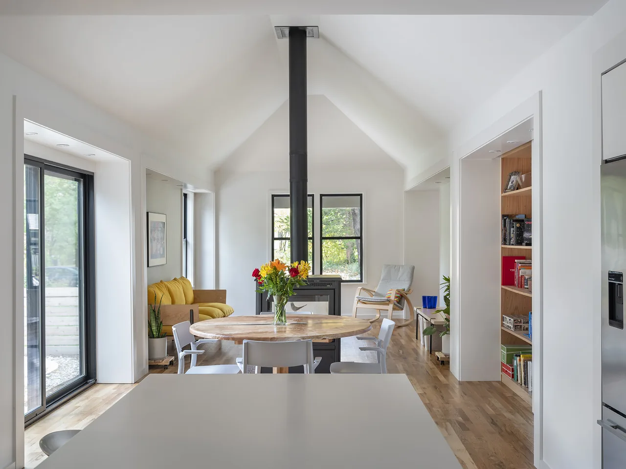 This open floor plan features a vaulted ceiling, central wood stove, and floor-to-ceiling glass doors, enhancing natural light and warmth.