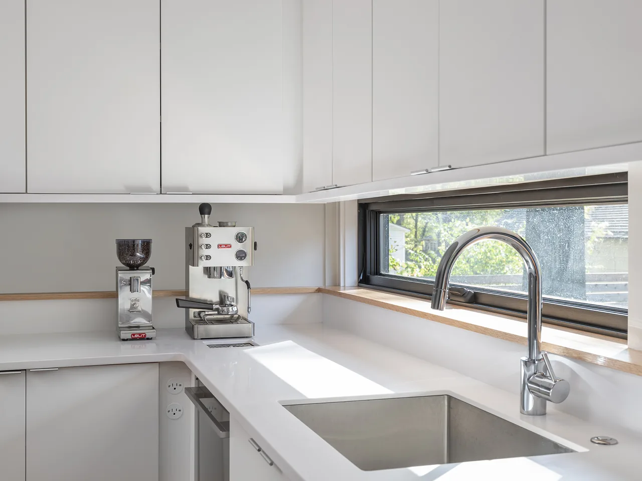 A modern kitchen corner featuring white cabinets, a built-in coffee machine, and a horizontal window above the stainless steel sink.