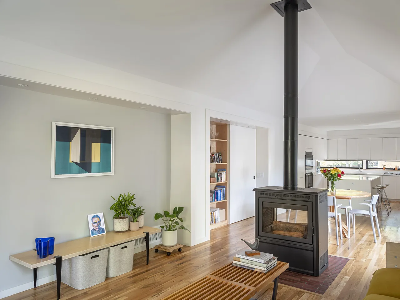 Open floor plan living area with central black freestanding wood stove and vaulted ceiling, leading to a white kitchen in the background.