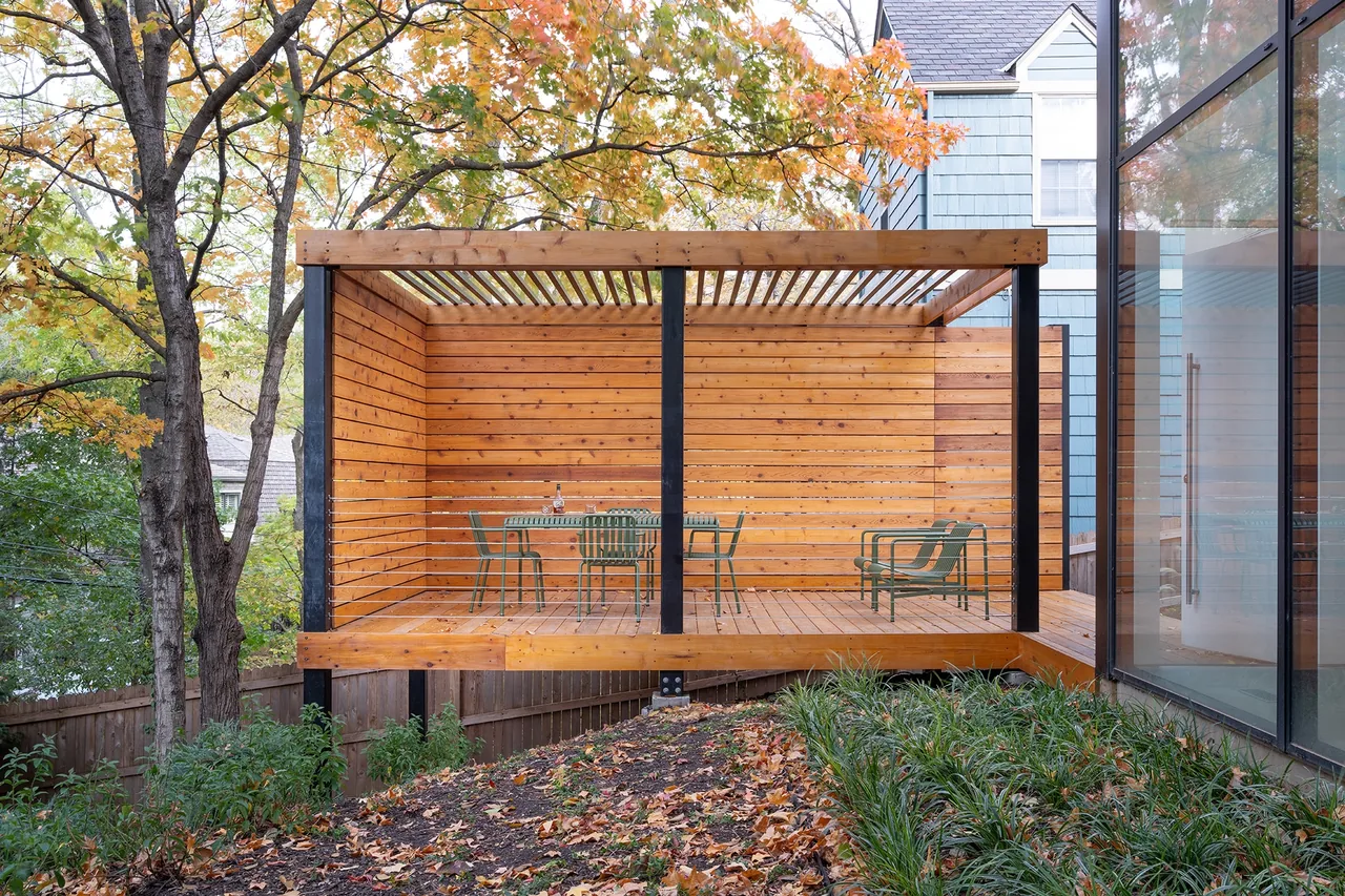 A wooden outdoor living patio with slatted roof and cable railing is set beside a modern house with large windows.