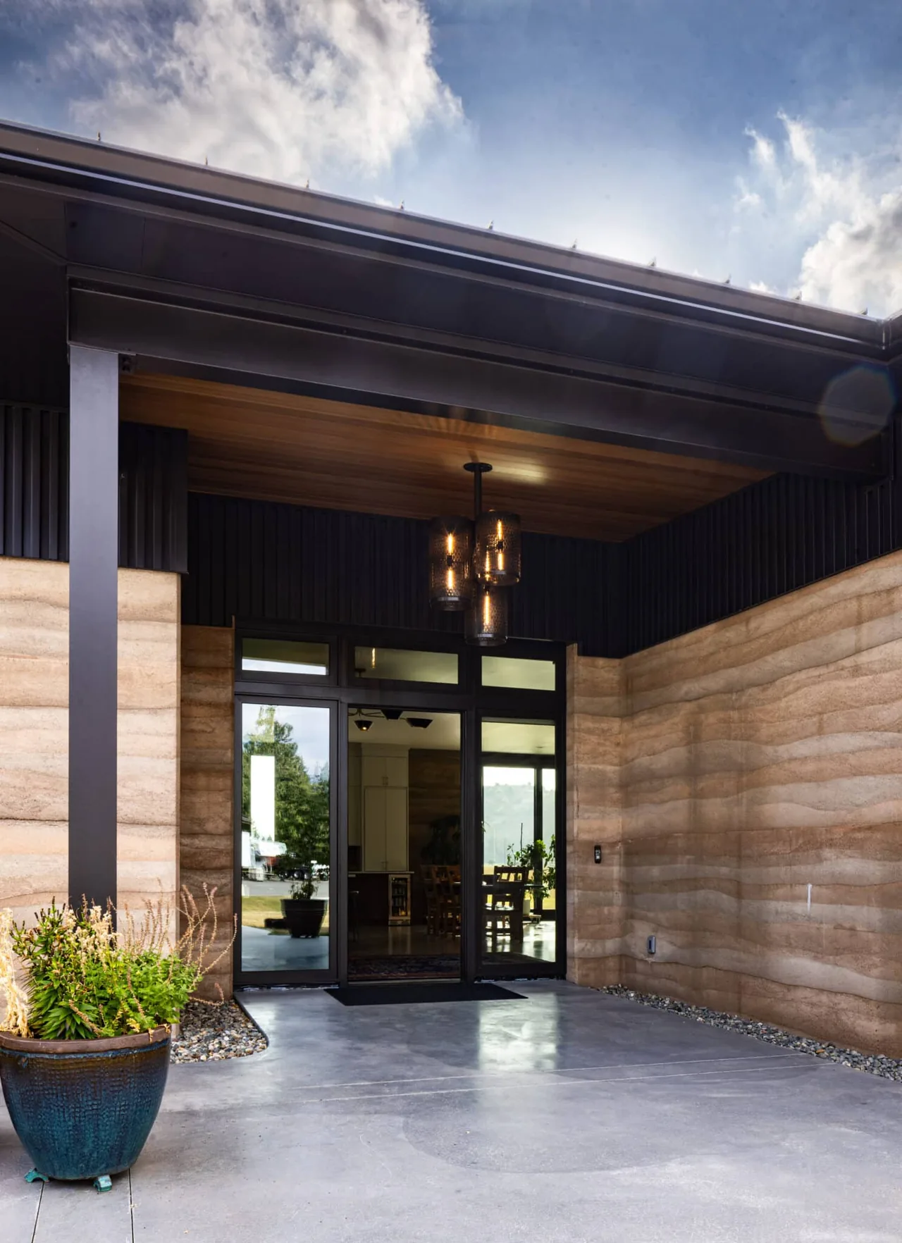 Entry of the Rammed Earth House with distinctive earth-tone walls, black window frames, and a wood ceiling with pendant lighting.