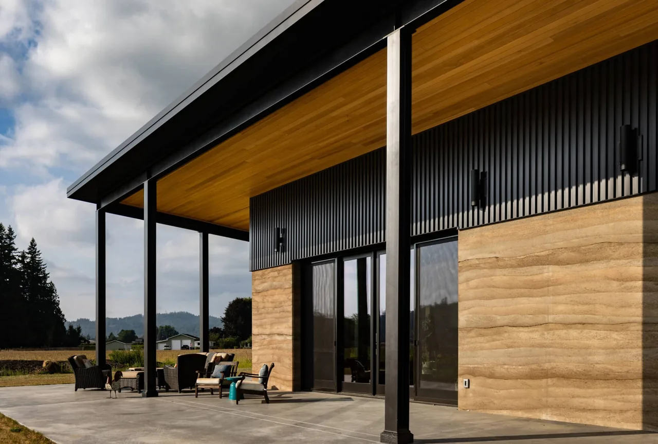Exterior view of a house with rammed earth walls and black steel beams supporting a wooden ceiling over the patio.