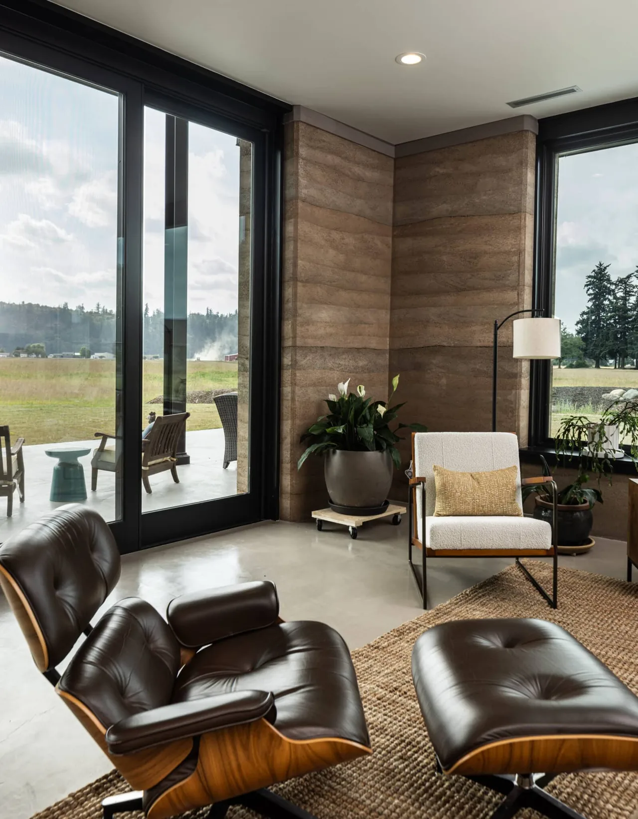 Living room corner features a leather lounge chair and ottoman with large black-framed windows and rammed earth walls.