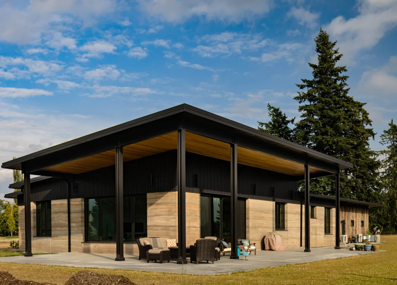 The Rammed Earth House features rammed earth walls, a black metal sloped roof, and an outdoor seating area under a covered patio.
