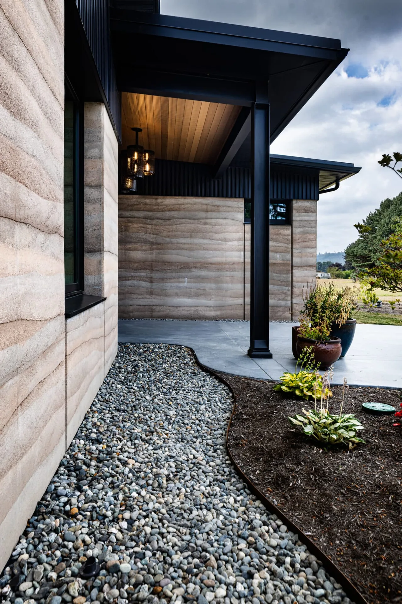 Rammed earth walls paired with black metal posts and a wood-paneled ceiling create a modern covered patio space.