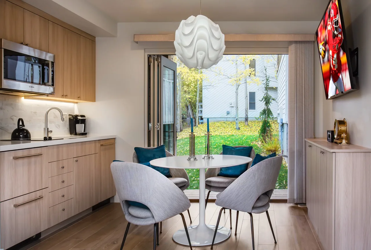Dining space adjacent to a kitchen with wood cabinetry and large folding glass doors overlooking a backyard with autumn leaves.