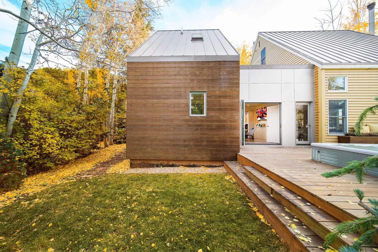Modern small house addition with wood siding, metal roofing, and a wooden deck featuring a hot tub, surrounded by autumn foliage.