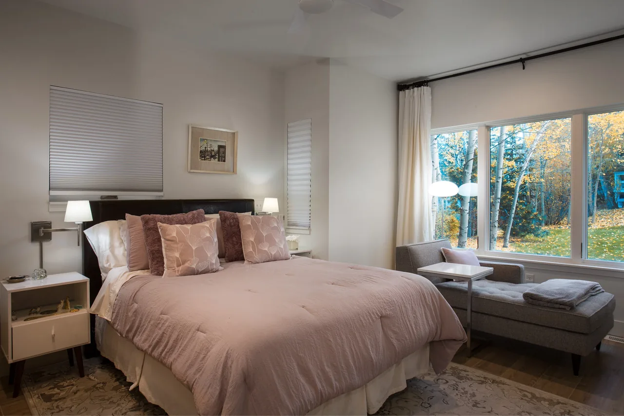 Bedroom featuring a large bed with pink bedding, two nightstands with lamps, and a lounge chair next to a big window overlooking autumn foliage.