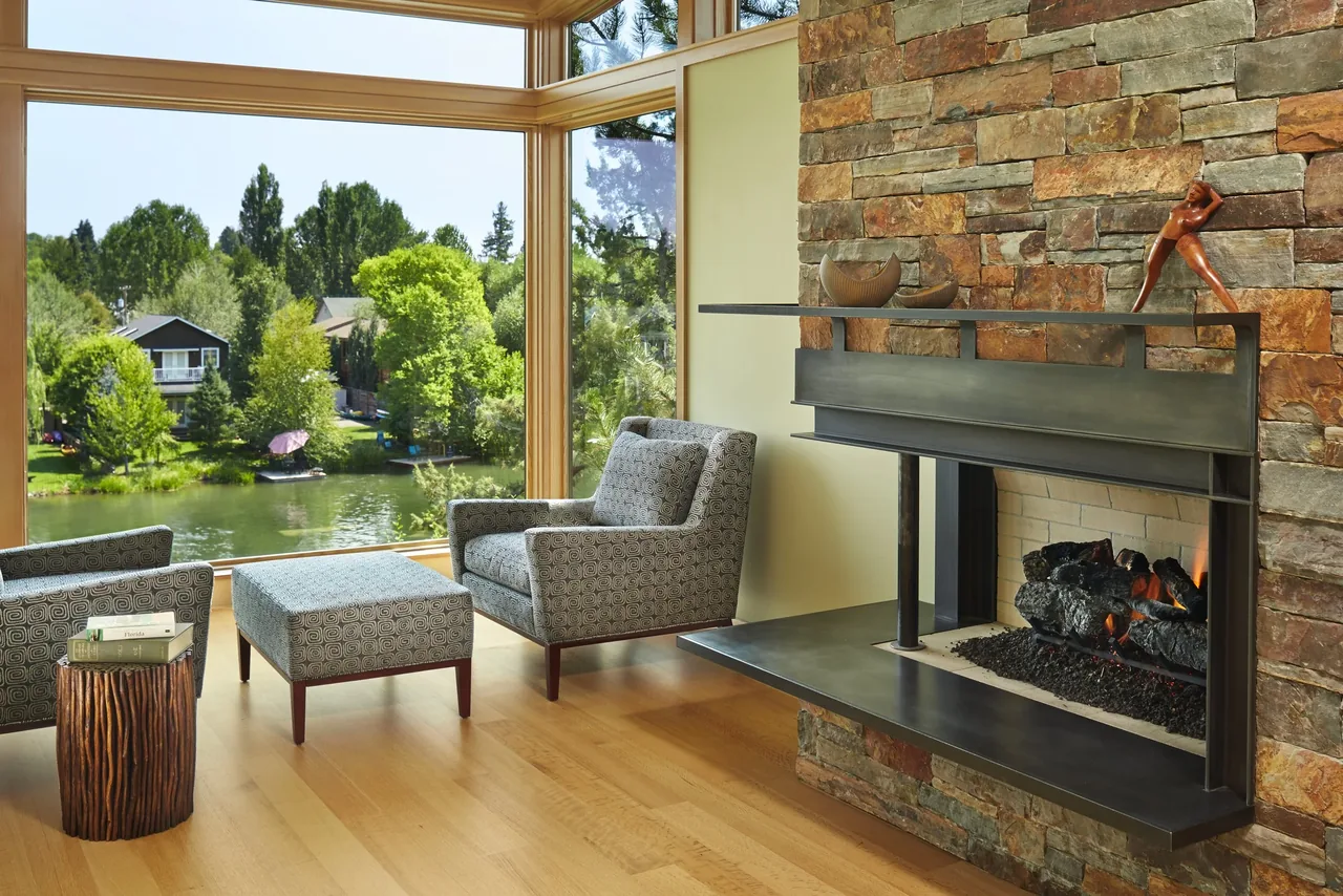 Living area features a stone fireplace and expansive wood-framed windows with lake views in the Deschutes project by FINNE Architects.