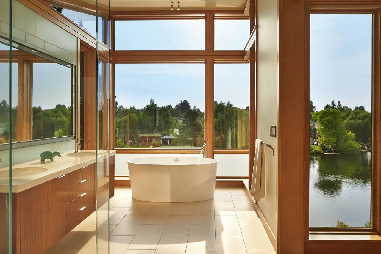 Modern bathroom features a freestanding tub by large wood window frames with lakefront scenery.
