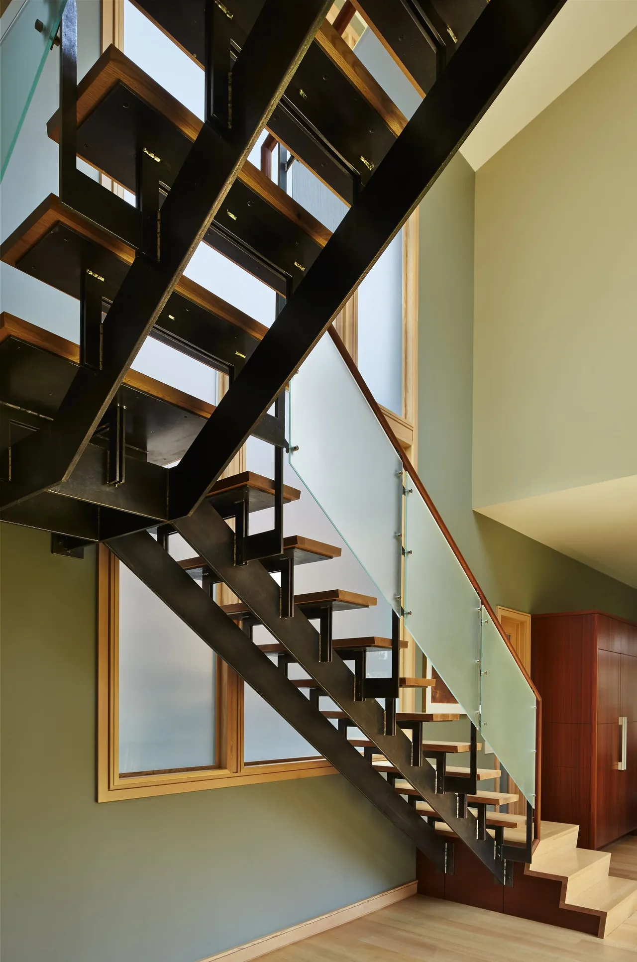 Interior view of a modern staircase featuring black steel stringers, wood treads, and a frosted glass railing with wood trim near large windows.