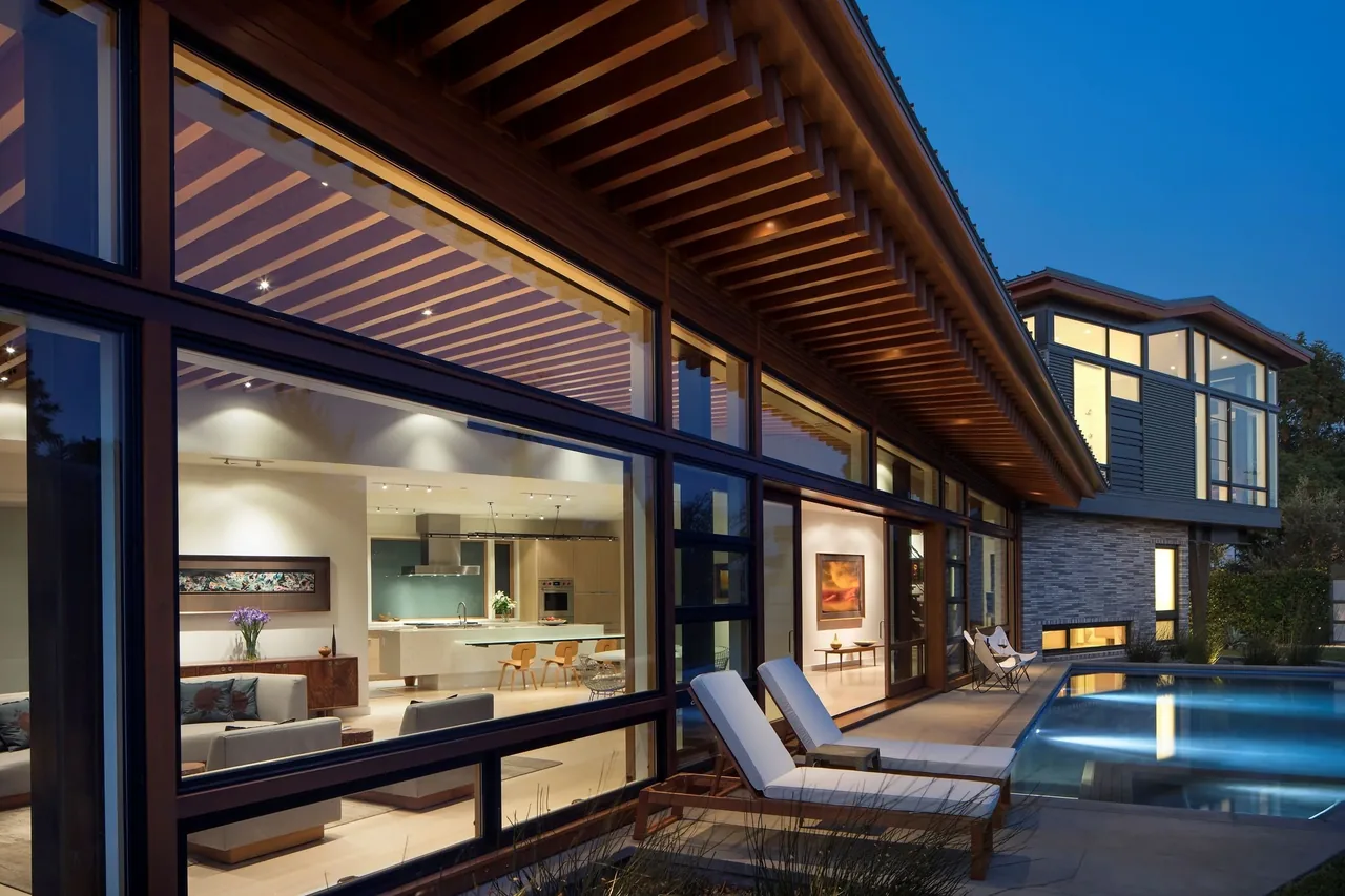 Contemporary home with extensive black window frames, covered patio, and narrow swimming pool at dusk by FINNE Architects.