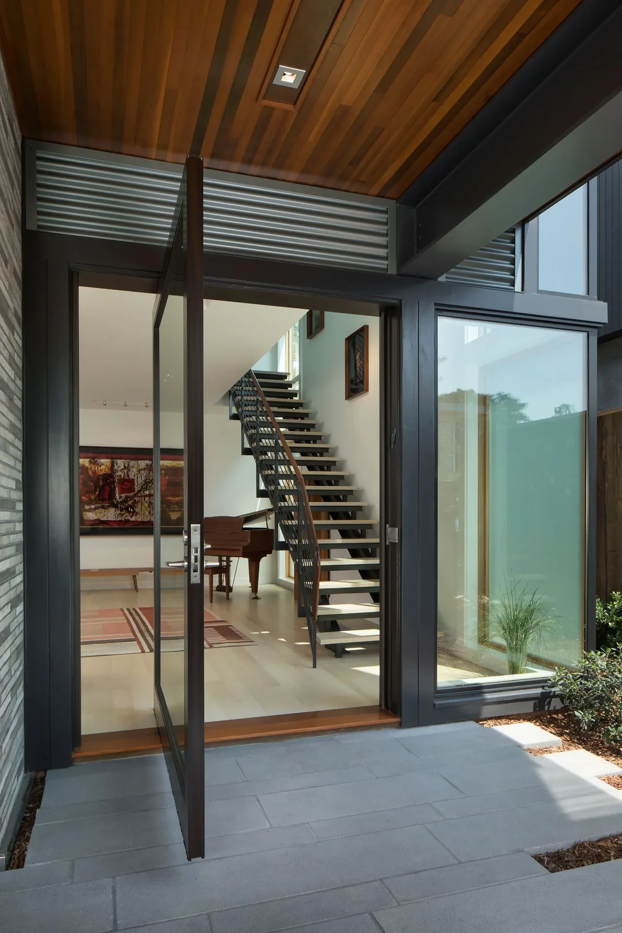 Open black-framed glass pivot door leads to a light-filled living room with floating stairs and piano.