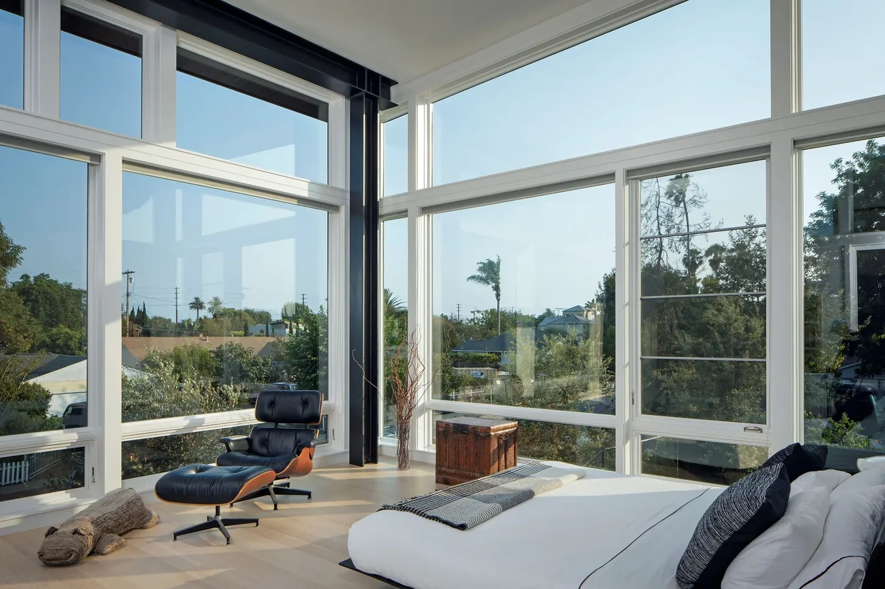 A bedroom corner features expansive floor-to-ceiling glazing with white frames overlooking greenery.
