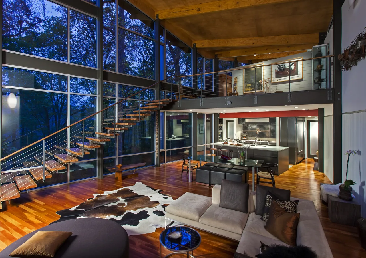 Open living area with large glass windows and floating stair connects to mezzanine above kitchen and dining space.