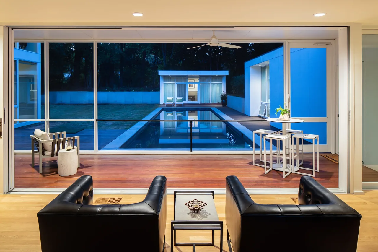 The living room features large sliding glass doors opening to a pool and a separate pool house in the evening.