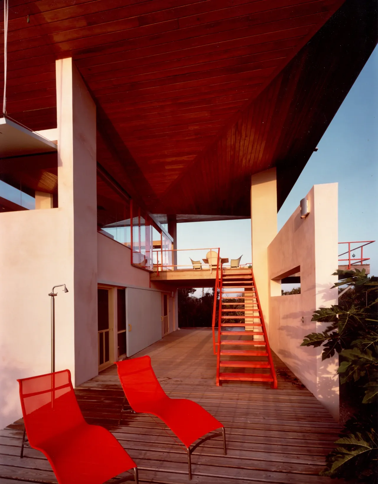 Taylor Vacation Home features a wooden deck terrace with modern red lounge chairs and a staircase under a large wood-paneled overhanging roof.