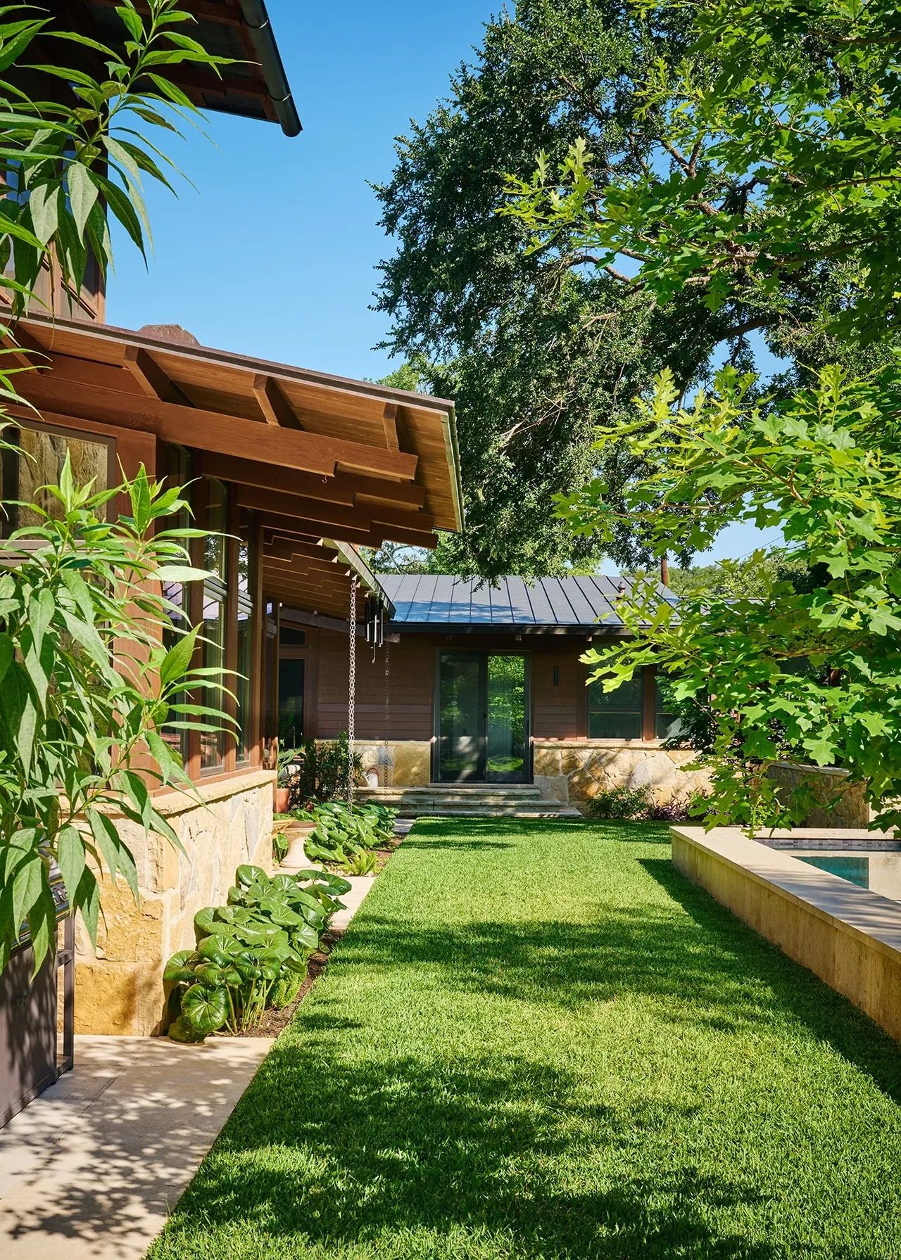 Modern residence with wood siding and natural stone base walls seen from a garden lawn with large shade trees.