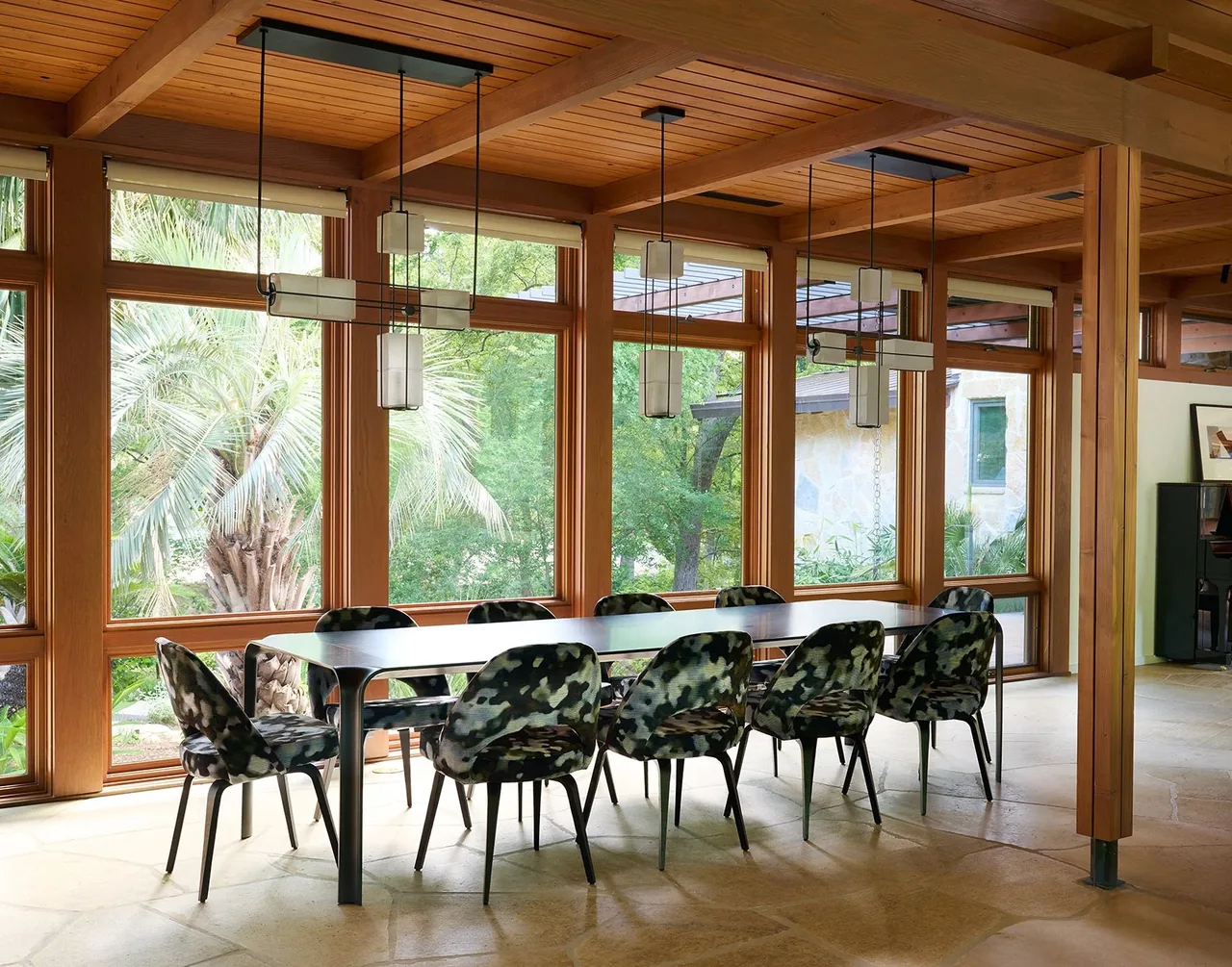 The dining area of FOOTHILL TERRACE showcases floor-to-ceiling wooden windows and pendant lighting with views of mature palm trees.