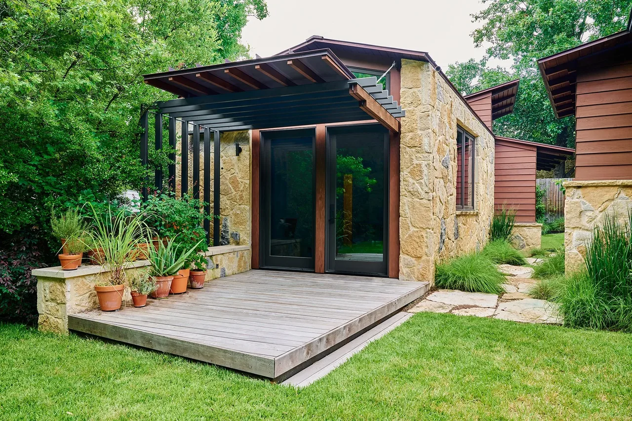 Exterior view of Foothill Terrace showing natural stone walls, a wooden deck, and black metal pergola with lush vegetation.