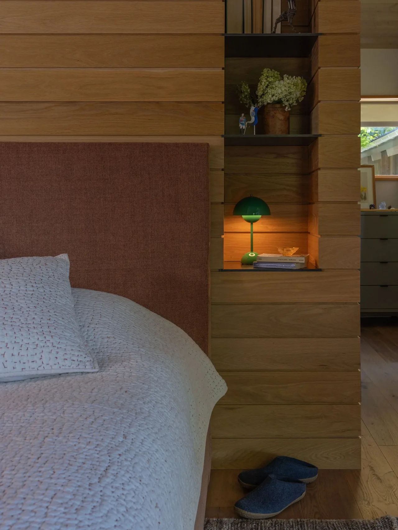 Wood-paneled bedroom wall with built-in shelves and a green lamp glowing beside a bed with an upholstered headboard.