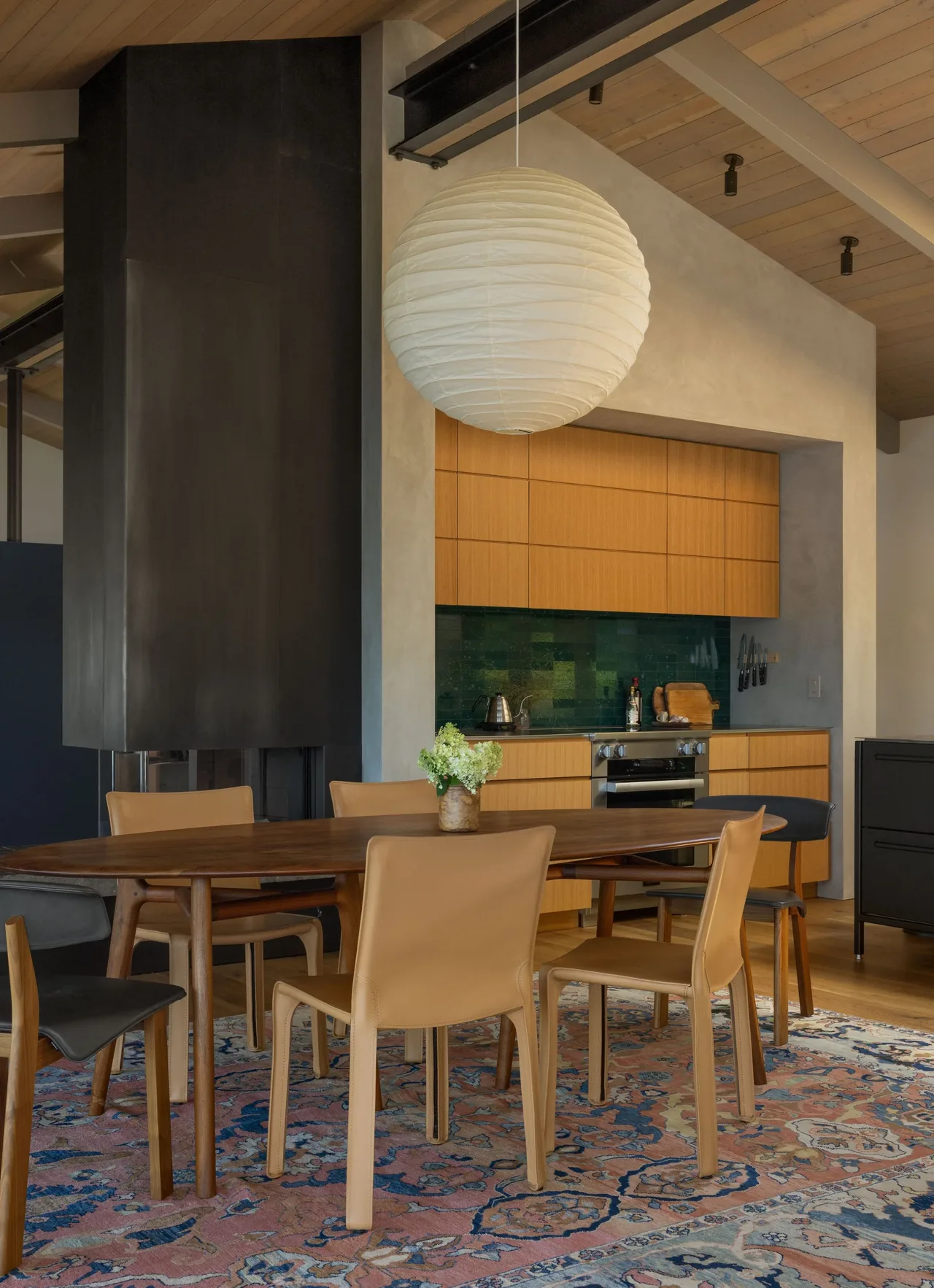 Dining space features a wood table with leather chairs and a paper lantern pendant light beside a modern black fireplace and wood kitchen cabinetry.