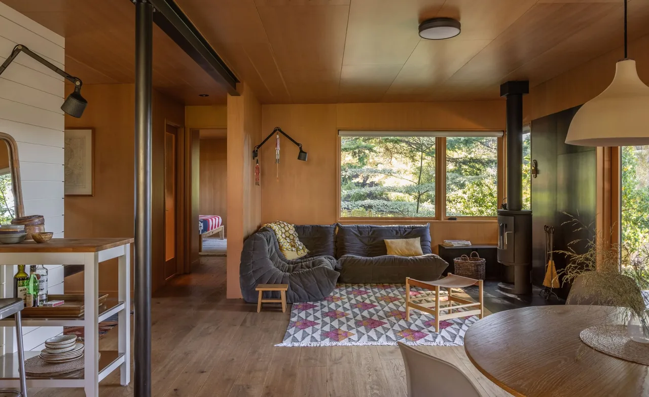 Living room with wood panel walls, a gray sectional sofa, and a wood stove by a large window in Eagle Harbor Residence.