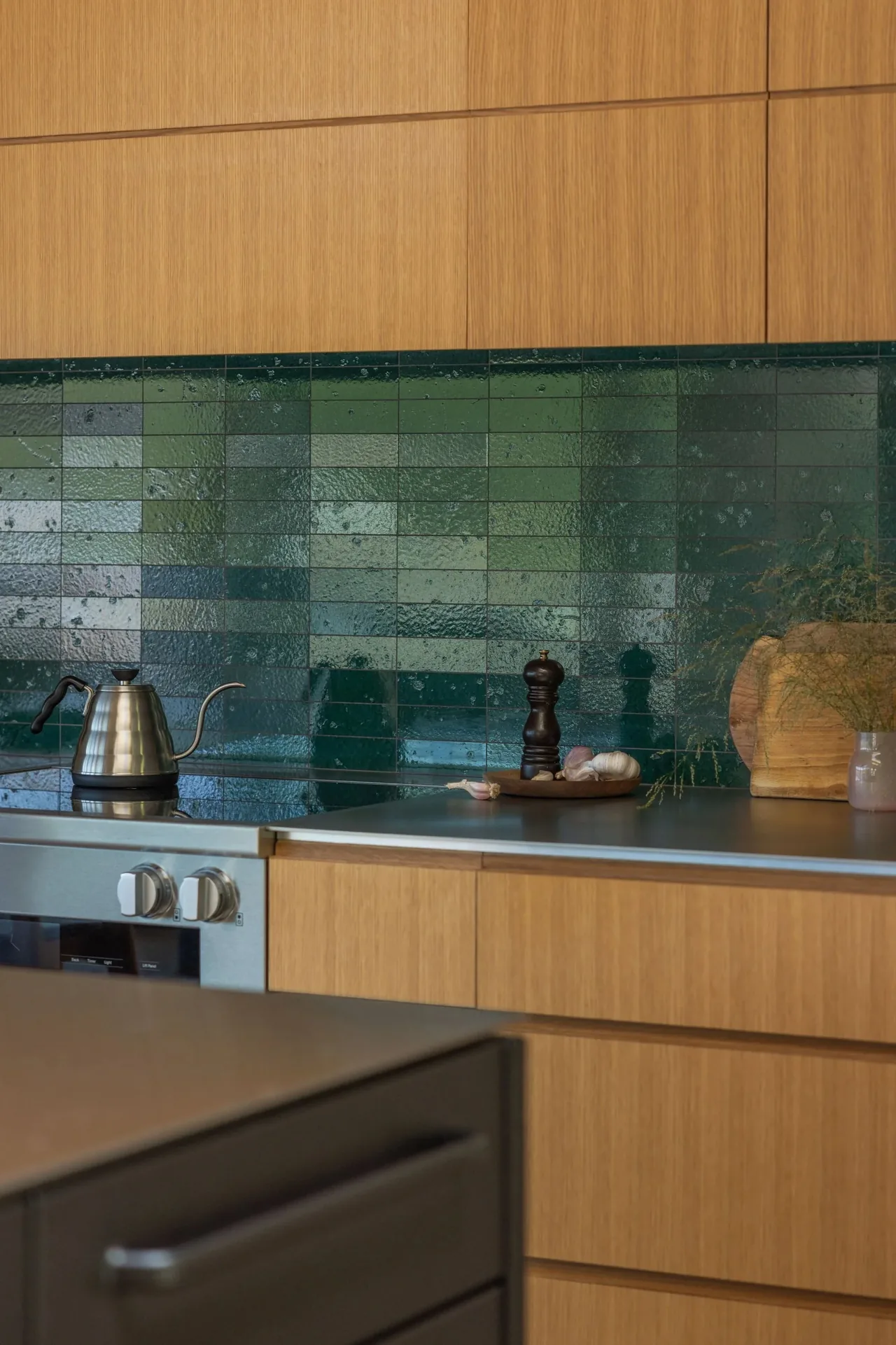 Kitchen features light wood cabinets, a glossy dark green tile backsplash, and a built-in stainless steel stove with control knobs.
