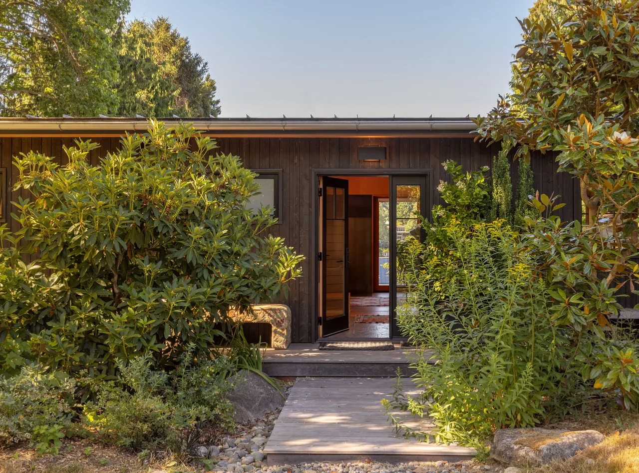 The Eagle Harbor Residence features a dark wood exterior with a flat roof and an entry door opening into a garden setting.