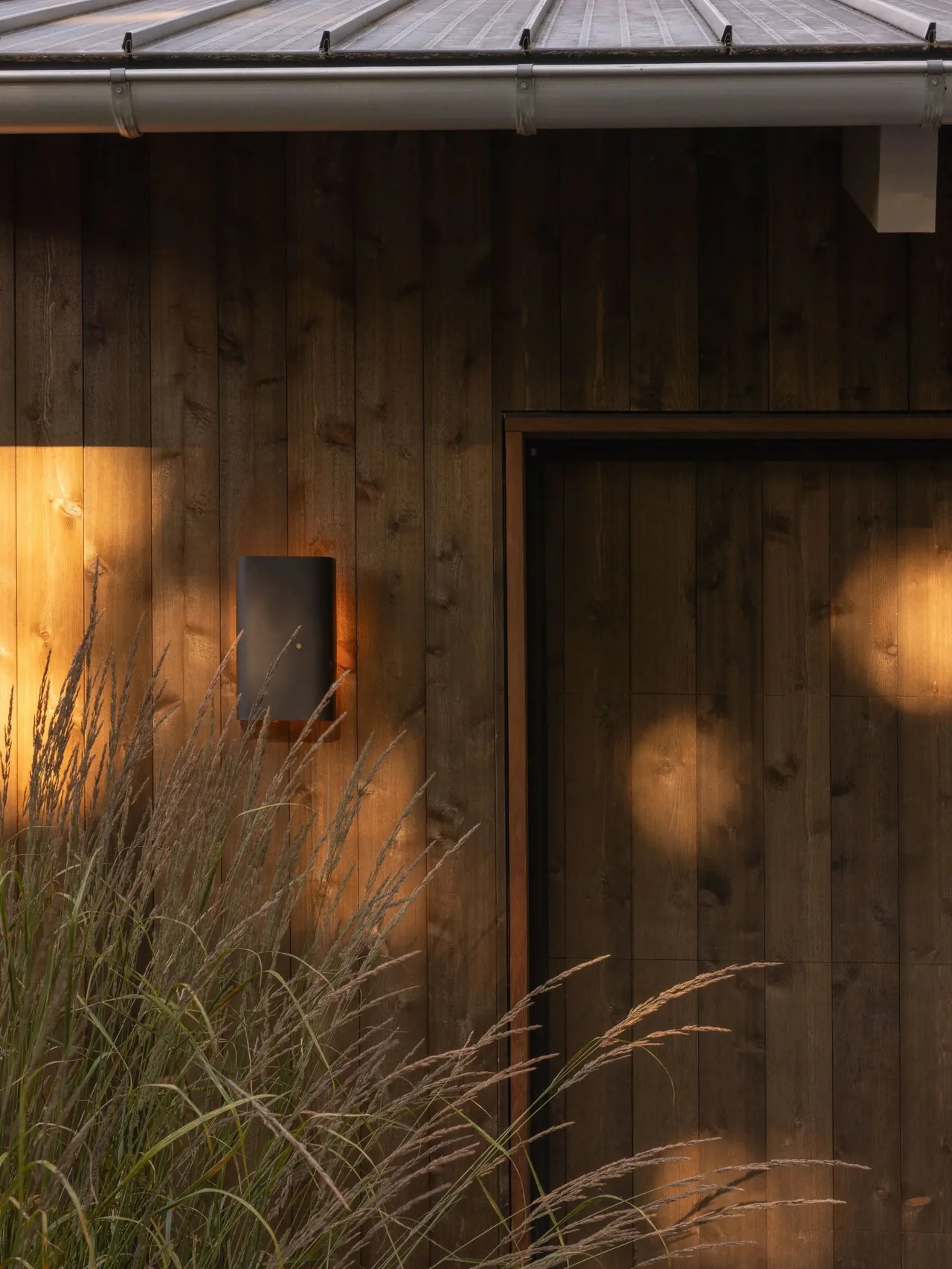 Exterior wood-paneled wall with integrated modern wall light and tall grasses at Eagle Harbor Residence.