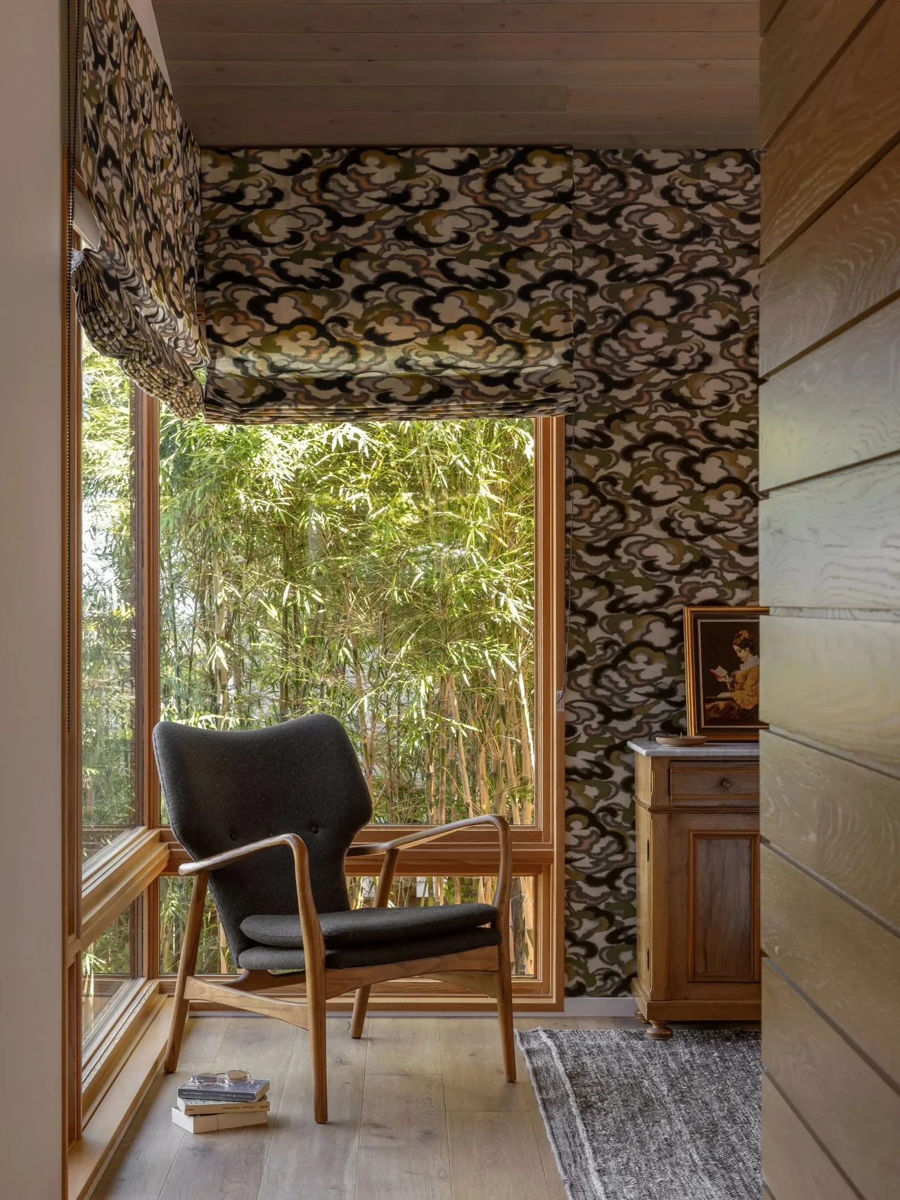 Corner seating area with a wood-framed chair next to floor-to-ceiling glazing and patterned Roman shades at Eagle Harbor Residence.