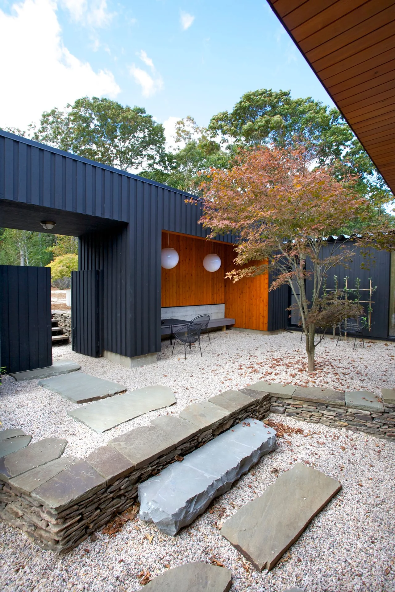 The Seaside House courtyard features gravel, stone seating walls, a stepping stone path, and a black wood-paneled exterior with a wood soffit.