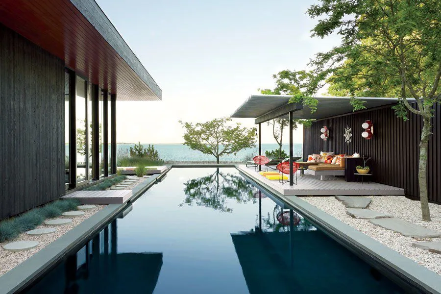 Seaside House features an outdoor pool adjacent to dark wood siding structures and a covered patio with seating, overlooking the water.