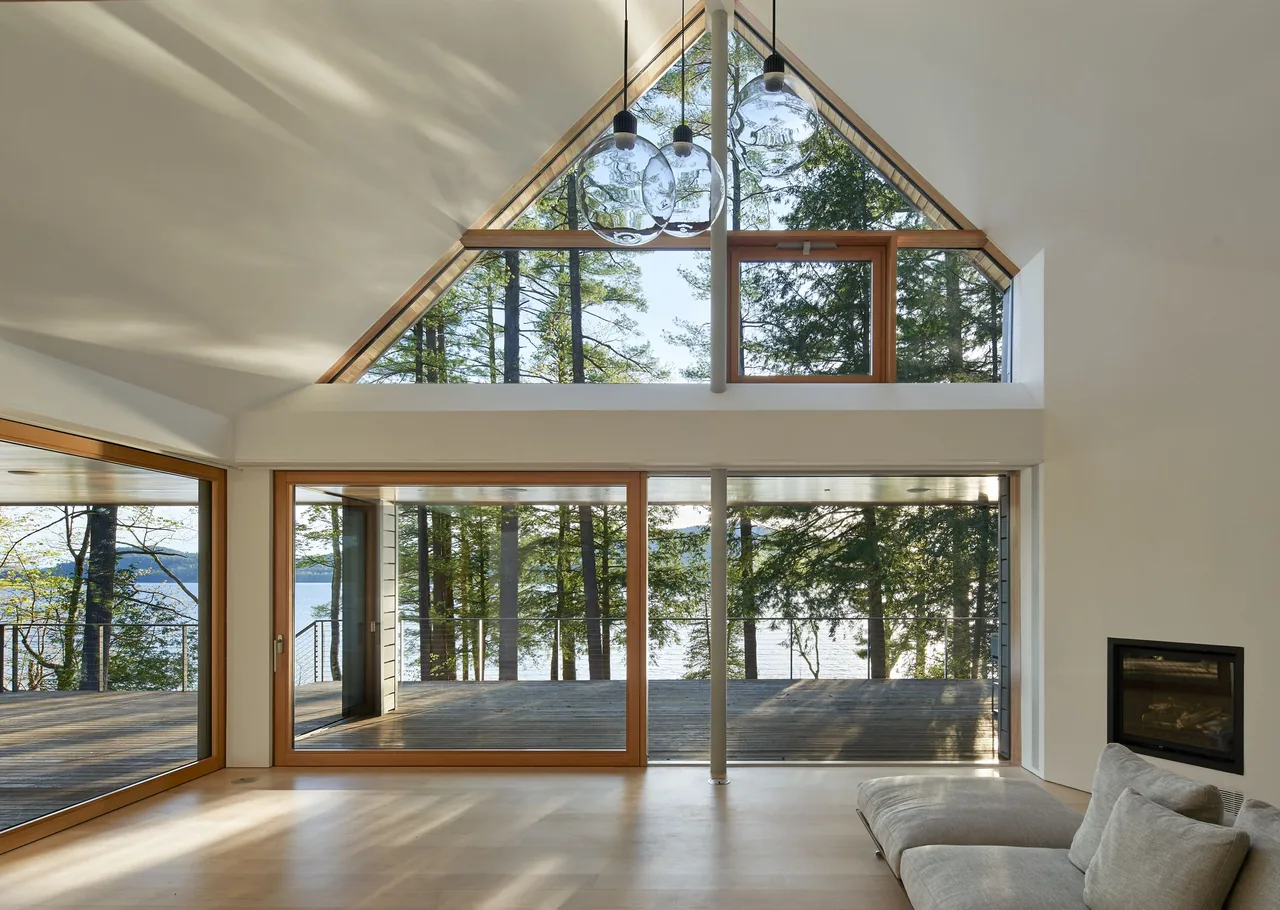 Living room with floor-to-ceiling wood-framed sliding glass doors and a triangular clerestory window overlooking a lakeside deck.