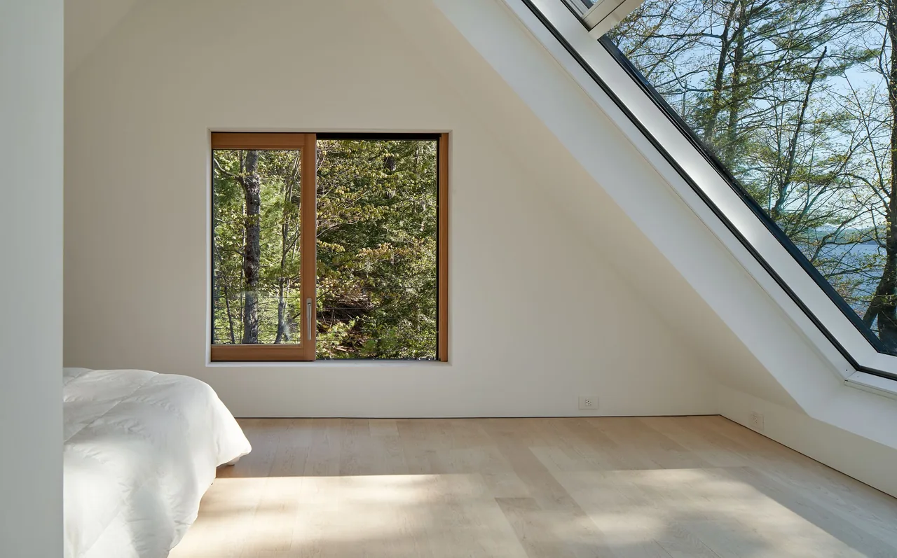 Bedroom with wood-framed window and large triangular glazed wall providing a forest view.