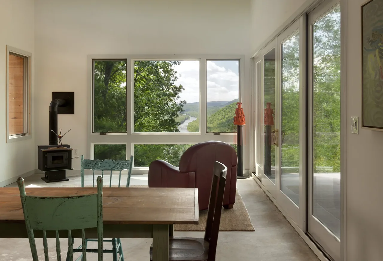 Dining area with wood stove and large windows framing a forested river view at Ridge House.
