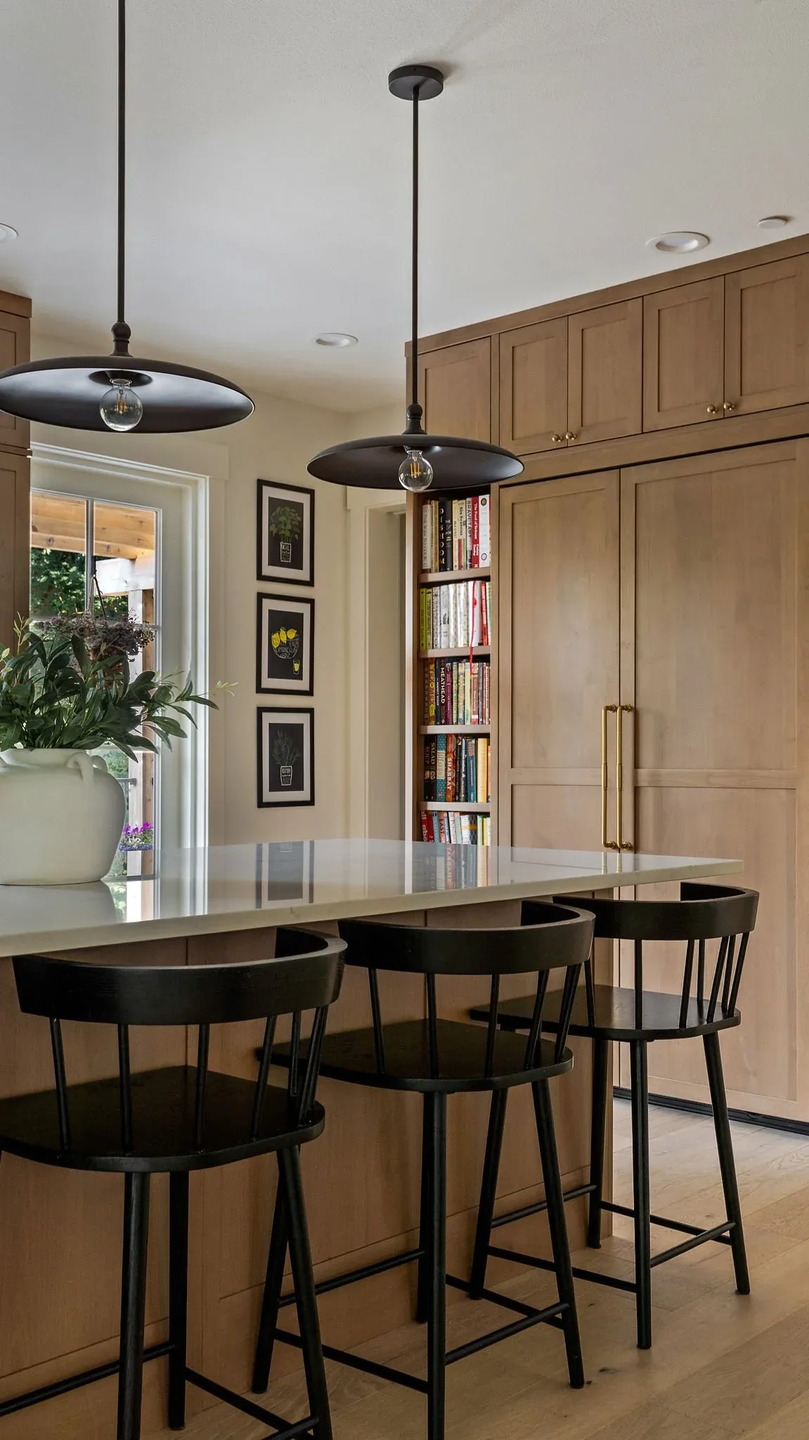 Kitchen island with white quartz countertop, black pendant lights, and black chairs, featuring light wood cabinets and a built-in bookcase.